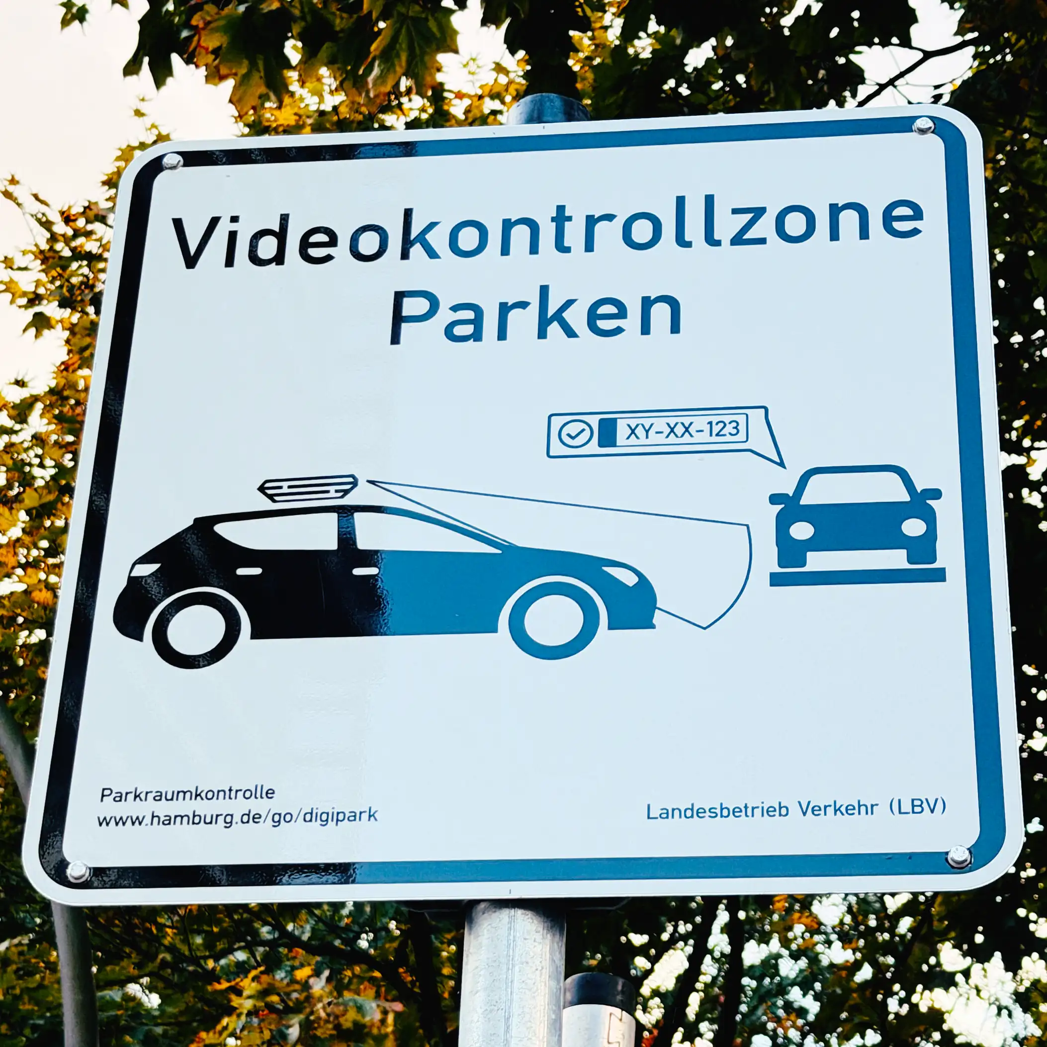 Das Bild zeigt ein Verkehrshinweisschild mit einer Silhouette eines Autos (Pkw) von der Seite. Auf dem Dach ist ein Kasten abgebildet, aus dem ein skizzierter Strahl oder Scan-Bereich auf die Fahrbahn scheint. Davor ist ein weiteres Auto abgebildet, das parkt. Ein fiktives Autokennzeichen XY-XX-123 wird dort angezeigt. Text über den beiden Fahrzeugen: "Videokontrollzone Parken". Am unteren Rand des Schildes steht: "Parkraumkontrolle. www.hamburg.de/go/digipark. Landesbetrieb Verkehr (LBV)".